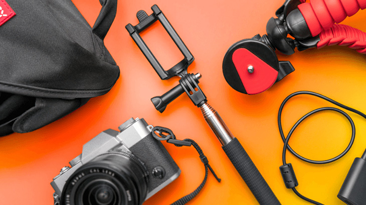 Na pomarańczowym tle leżą: aparat, torba, kijek do selfie, statyw z czerwonymi nogami i czarny kabel.