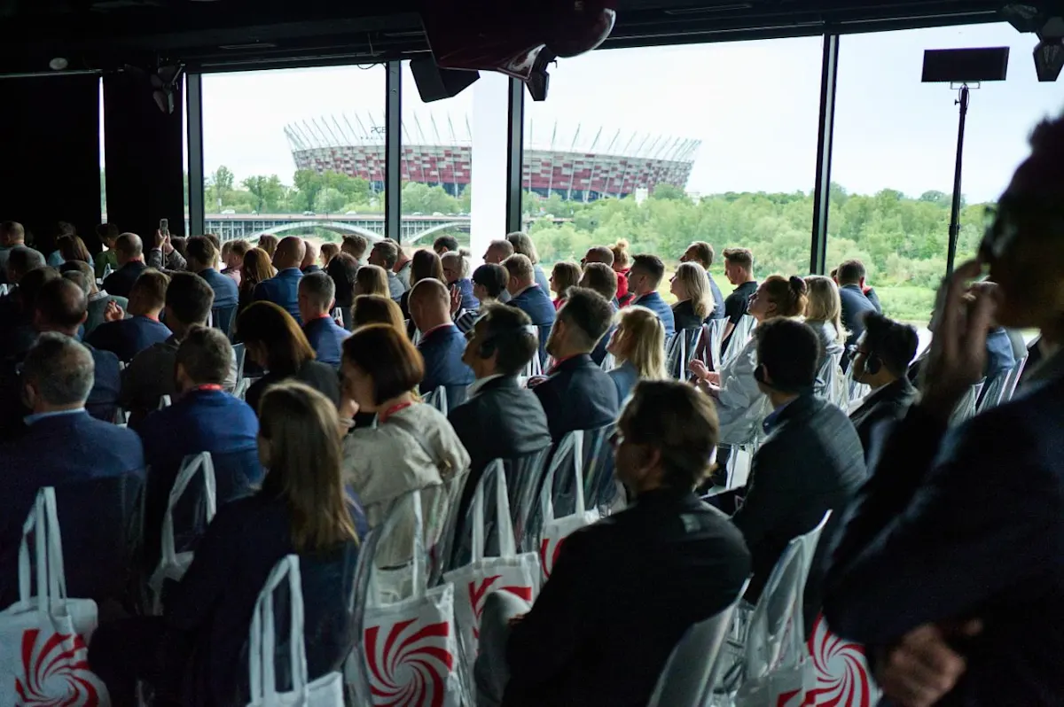 Widok na salę konferencyjną z siedzącymi ludźmi. W tle okna z widokiem na Stadion Narodowy i zieleń. Torby leżą na krzesłach.