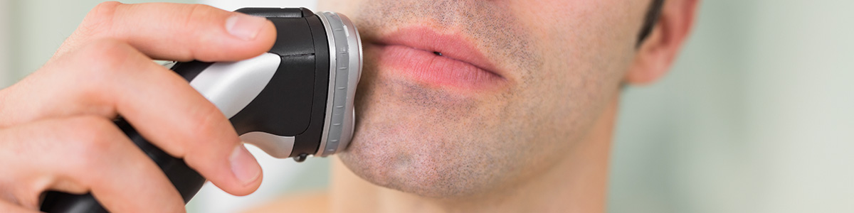 Mężczyzna goli się elektryczną golarką. Golarka jest czarno-srebrna, a dłoń trzyma ją przy twarzy. Tło jest jasne.