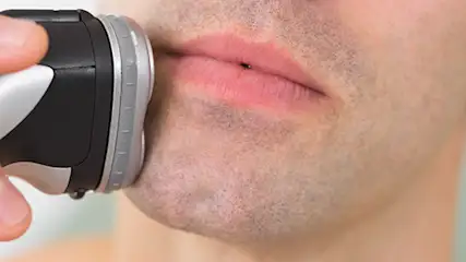 Mężczyzna goli się elektryczną golarką. Golarka jest czarno-srebrna, a dłoń trzyma ją przy twarzy. Tło jest jasne.