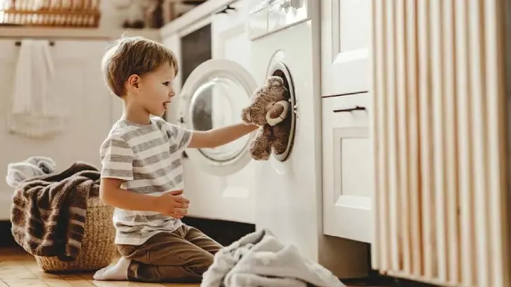 Chłopiec wkłada pluszowego misia do pralki. Obok kosz z praniem i białe ubrania na podłodze. W tle meble.