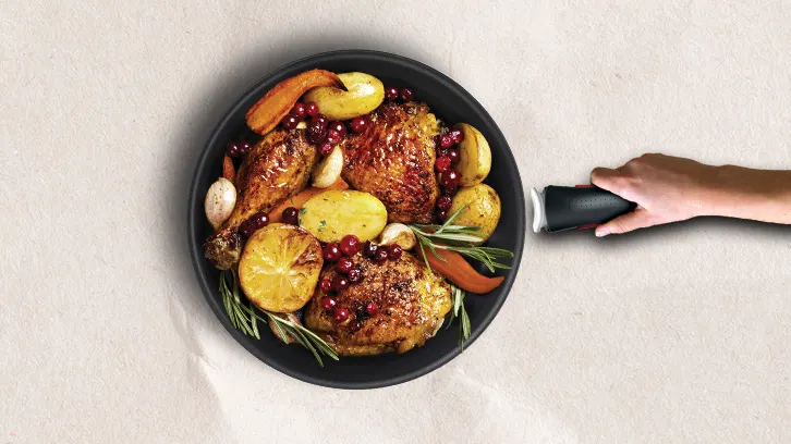 Czarny rondel z pieczonym kurczakiem, ziemniakami, marchewką, czosnkiem, rozmarynem i żurawiną, trzymany za uchwyt.