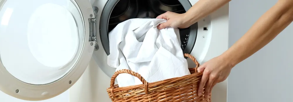 Otwarty, biały bęben pralki z ubraniami i kosz wiklinowy trzymany przez dwie ręce.