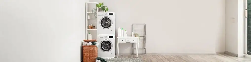 Zdjęcie przedstawia pralnię z białą pralką i suszarką, drewnianym koszem na pranie i białym stolikiem. W tle białe ściany i drewniana podłoga.
