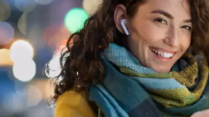 Uśmiechnięta kobieta z słuchawką w uchu trzyma telefon. Ma na sobie żółtą kurtkę i szal w odcieniach zieleni i niebieskiego. W tle światła miasta.