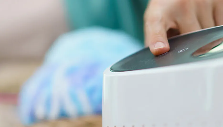 Dłoń naciska przycisk na białym urządzeniu. W tle widoczna osoba w zielonej bluzce i niebieski materiał.