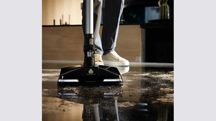 Osoba czyści podłogę mopem elektrycznym. Czarny mop ze srebrną rączką stoi na mokrej, brązowej podłodze. W tle widać nogi osoby w szarych spodniach.