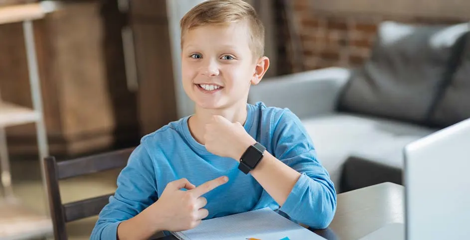 Uśmiechnięty chłopiec w niebieskiej koszulce wskazuje na czarny smartwatch na nadgarstku, siedząc przy biurku z laptopem i notatnikiem.