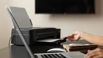 Kobieta przy biurku z laptopem i drukarką, wyjmuje kartkę papieru. W tle telewizor na ścianie.