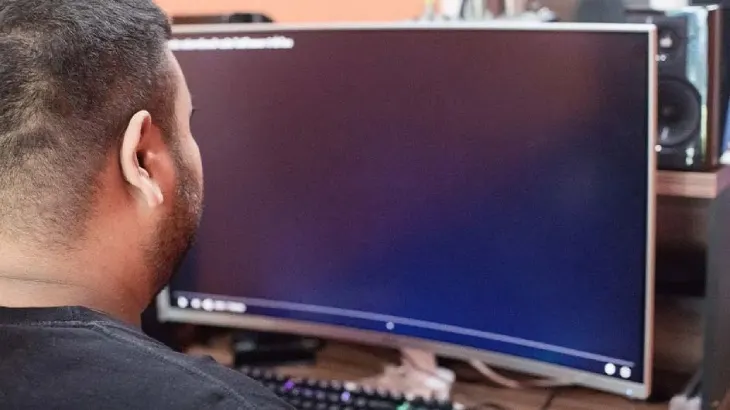 Mężczyzna siedzi przed zakrzywionym monitorem i klawiaturą. W tle widoczne głośniki i drewniany blat biurka.