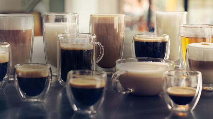 Na zdjęciu widoczne są różne szklanki i filiżanki z kawą, mlekiem i innymi napojami na ciemnym blacie.