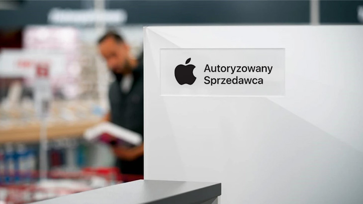 Biały znak z czarnym logo Apple i napisem "Autoryzowany Sprzedawca". W tle zamazana sylwetka osoby w sklepie.
