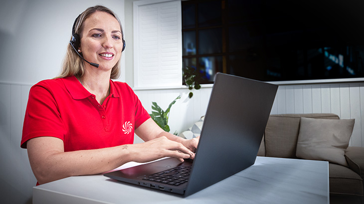 Kobieta w czerwonej koszulce z zestawem słuchawkowym używa laptopa na biurku. Za nią widać roślinę i kanapę.