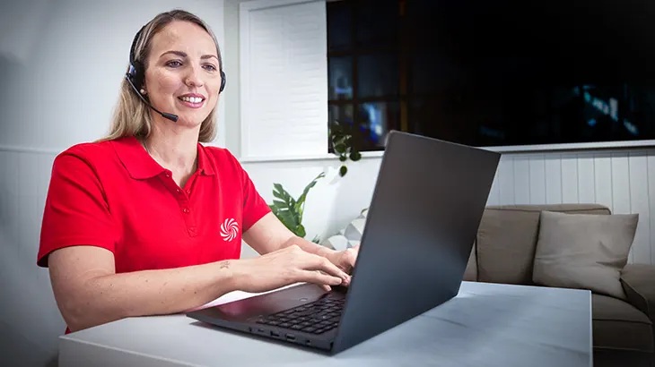 Kobieta w czerwonej koszulce z zestawem słuchawkowym używa laptopa na biurku. Za nią widać roślinę i kanapę.