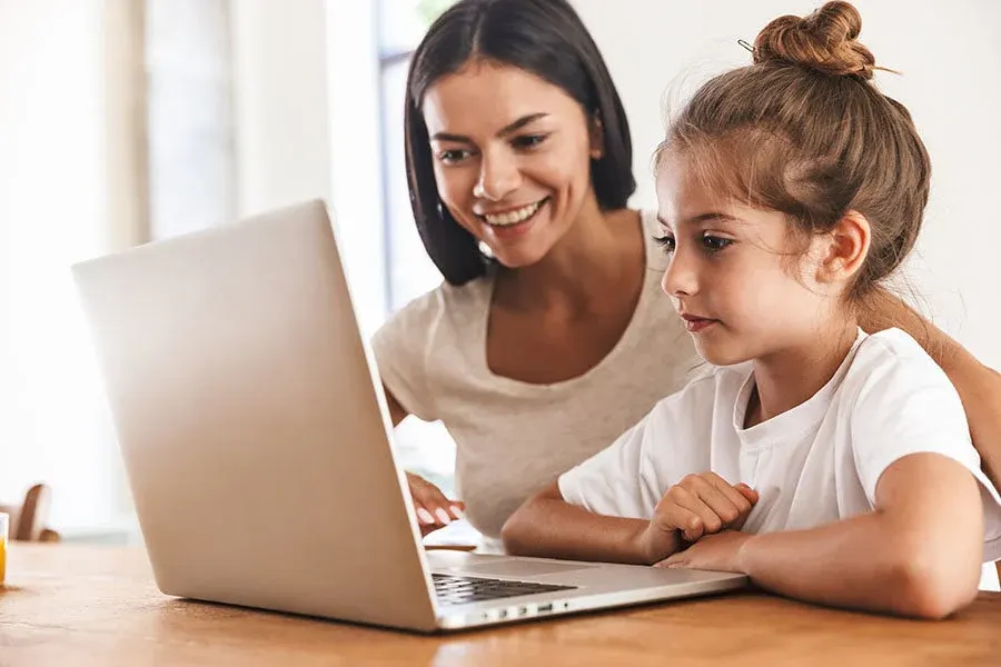 Mama i córka patrzą na laptopa na drewnianym stole. Obie ubrane są w białe koszulki. W tle widać okno.