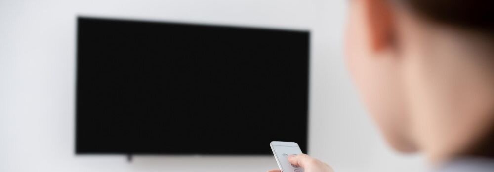 Osoba trzyma biały pilot przed czarnym telewizorem na białej ścianie.