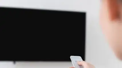 Osoba trzyma biały pilot przed czarnym telewizorem na białej ścianie.