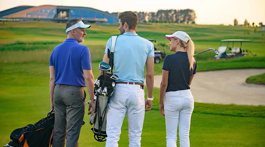Trzy osoby na polu golfowym: mężczyzna z przodu z torbą, mężczyzna z boku i kobieta z kijem golfowym. W tle widać wózek golfowy.
