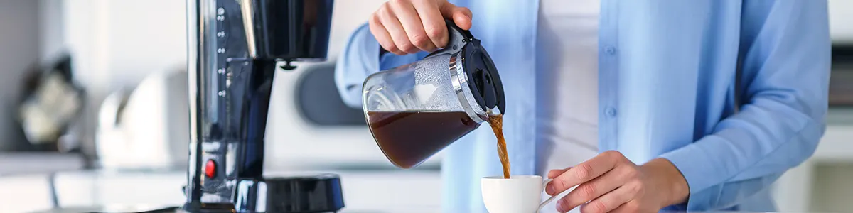 Osoba w niebieskiej koszuli nalewa kawę z dzbanka do białego kubka obok czarnego ekspresu do kawy.