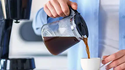 Osoba w niebieskiej koszuli nalewa kawę z dzbanka do białego kubka obok czarnego ekspresu do kawy.