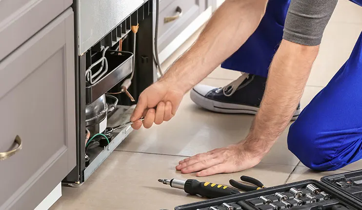 Mężczyzna w niebieskim kombinezonie naprawia lodówkę, używając klucza. Obok leżą narzędzia w czarnej skrzynce.