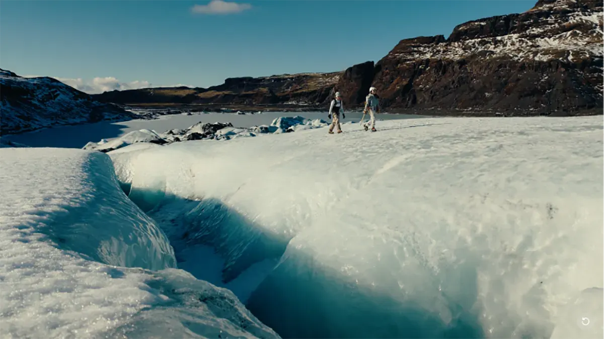 Dwóch turystów idzie po lodowcu; na pierwszym planie głęboka szczelina lodowa, w tle ciemne, ośnieżone zbocza i błękitne niebo.