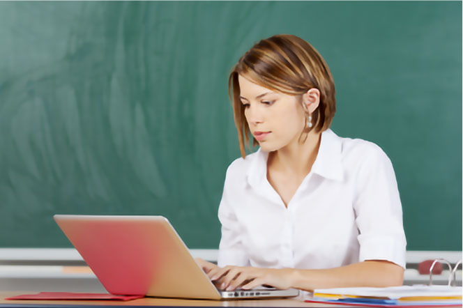 Kobieta w białej koszuli używa laptopa przy biurku. W tle zielona tablica i dokumenty.