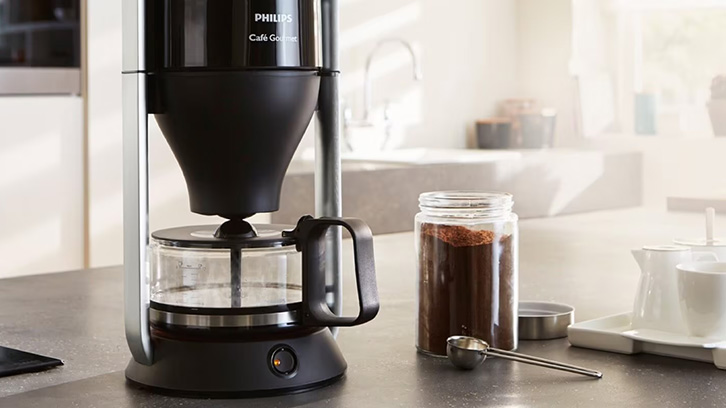 Czarny ekspres do kawy na blacie obok słoika z kawą i łyżeczki. W tle białe meble.