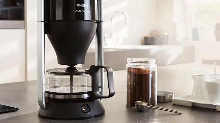 Czarny ekspres do kawy na blacie obok słoika z kawą i łyżeczki. W tle białe meble.