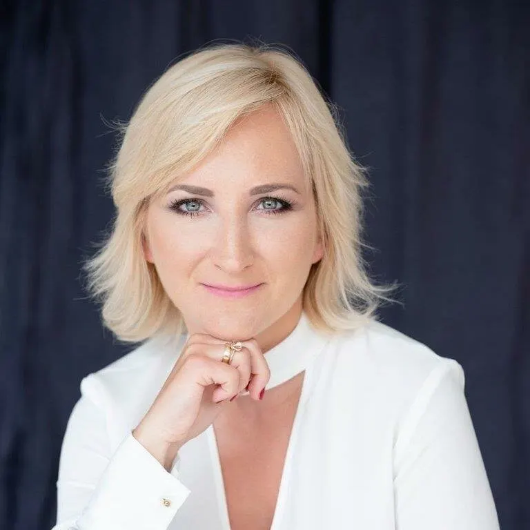 Kobieta w białym żakiecie opiera brodę na dłoni z pierścionkiem, tle jest ciemna zasłona.