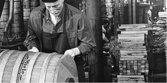 Czarno-białe zdjęcie przedstawia pracownika w fartuchu, który pracuje nad drewnianą beczką z napisem "Miele" w fabryce. W tle widać stosy drewna.