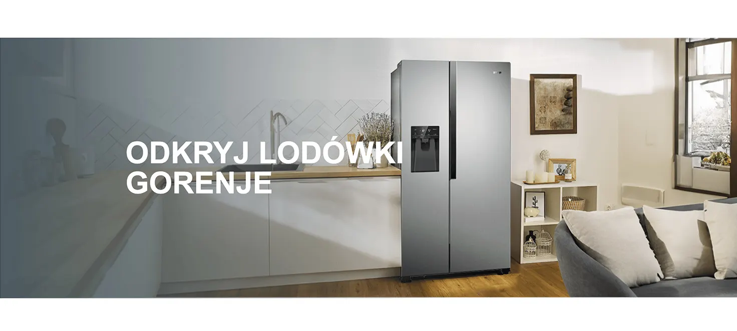 Lodówka Gorenje, dwudrzwiowa, srebrna, stoi w kuchni obok blatu i szafek. W tle okno i sofa.
