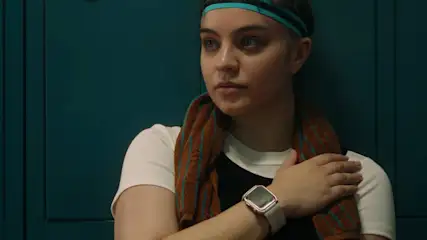 Młoda osoba z opaską na głowie i ręcznikiem na ramionach stoi na tle szatni z metalowymi szafkami.