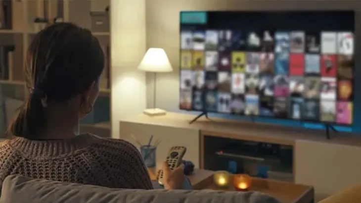 Kobieta z tyłu siedzi na kanapie z pilotem. Przed nią telewizor z wieloma miniaturami. Obok lampka i świece.
