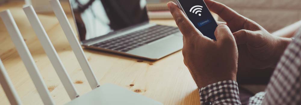 Osoba trzyma telefon z ikoną Wi-Fi obok routera i laptopa na drewnianym stole.