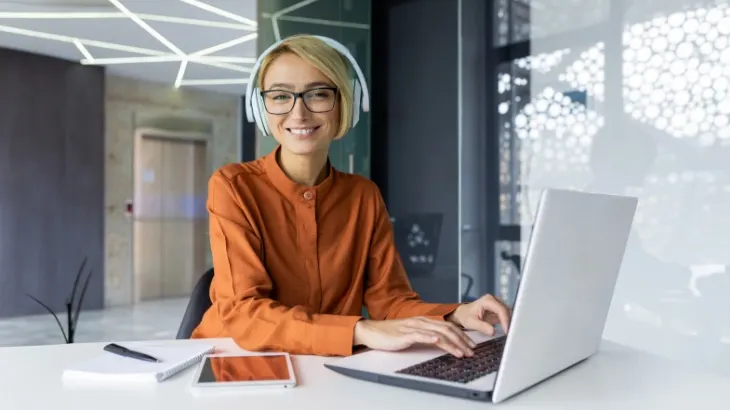 Kobieta w słuchawkach i okularach pracuje na laptopie przy biurku. Obok leżą notatnik i tablet. W tle widoczne okno i dekoracja sufitu.