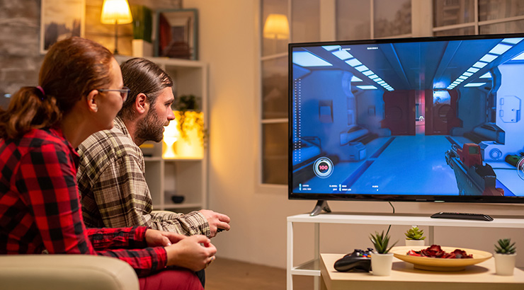 Dwoje ludzi siedzi na kanapie, grając w grę wideo na dużym telewizorze. Na stoliku pod telewizorem leżą przekąski i kontroler.