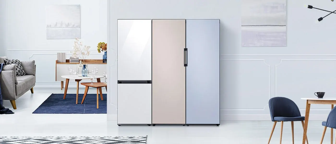 Wnętrze pokoju z lodówką o trzech kolorowych drzwiach: białym, beżowym i błękitnym. W tle meble i dekoracje w jasnych tonacjach.