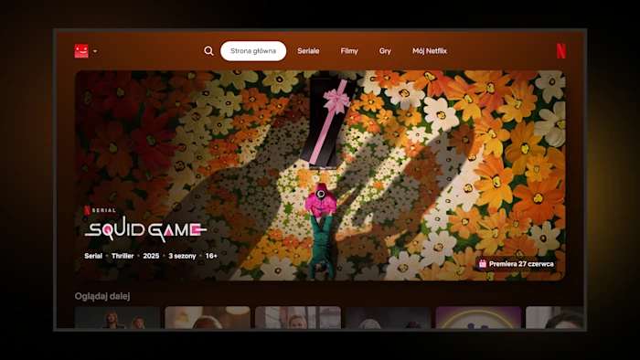 Interfejs aplikacji Netflix na telewizorze wyświetlający zapowiedź serialu "Squid Game". Na ekranie widać postać w zielonym dresie leżącą na kwiatowej posadzce obok czarnej trumny z różową wstążką.