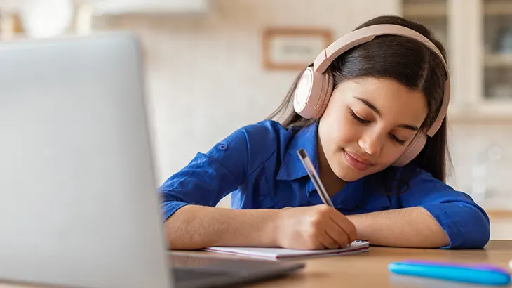 Dziewczyna w słuchawkach pisze w notatniku przy laptopie. Na stole leży długopis i etui. Tło rozmyte, jasne.