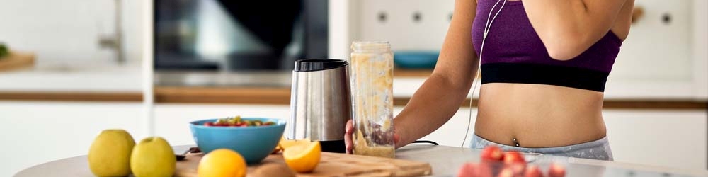 Kobieta w staniku sportowym przygotowuje koktajl w kuchni. Na stole owoce, blender i miska z jedzeniem.