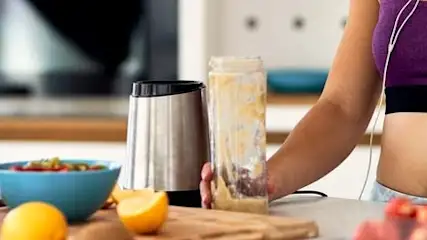 Kobieta w staniku sportowym przygotowuje koktajl w kuchni. Na stole owoce, blender i miska z jedzeniem.