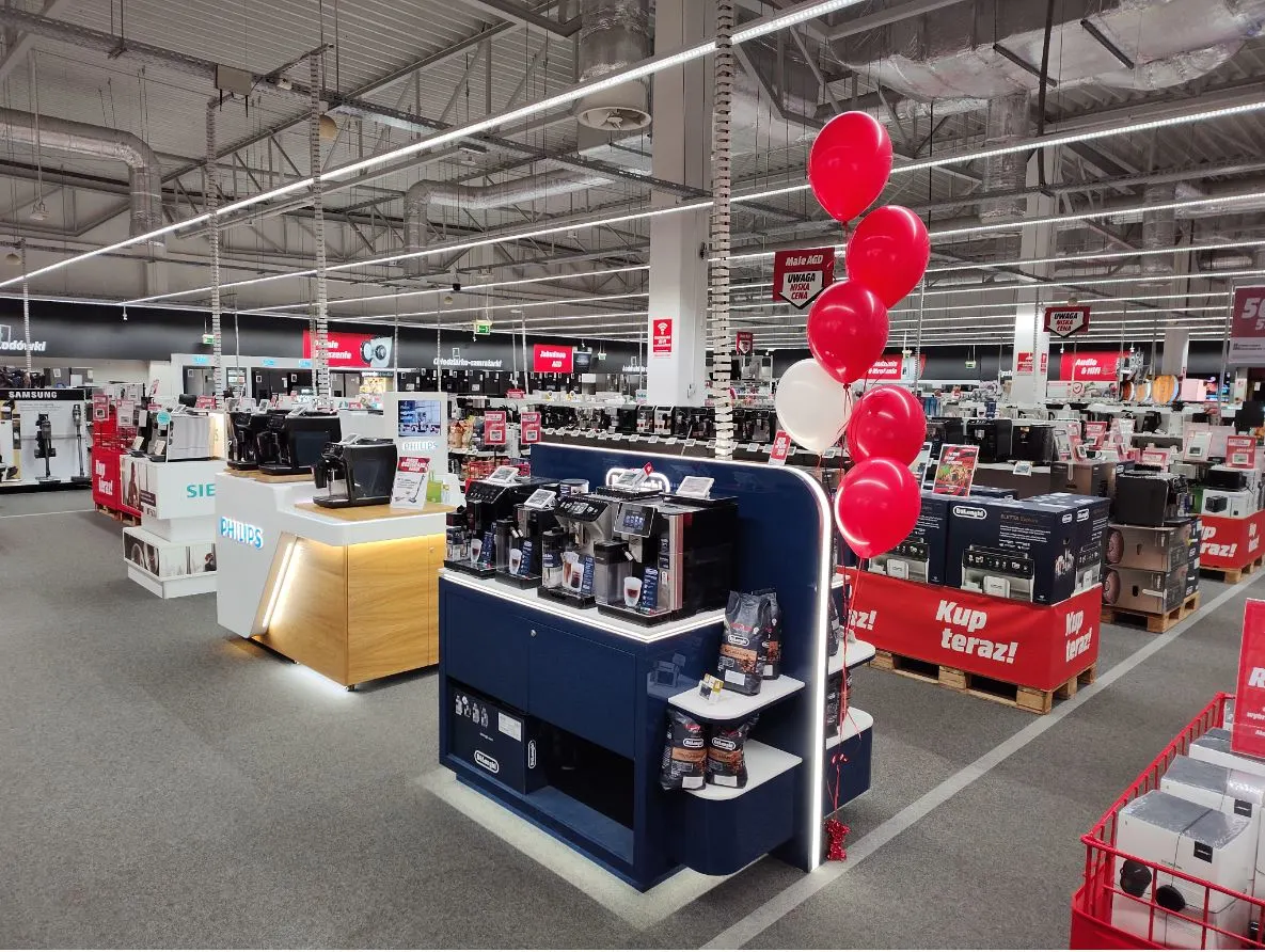 Wnętrze sklepu z ekspozycją ekspresów do kawy. Czerwone i białe balony unoszą się nad stoiskami. Plakaty promocyjne widoczne w tle.