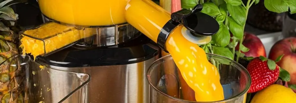 Sokowirówka robi sok, w tle owoce: ananas, jabłka, truskawka, cytryna i bazylia. Żółty sok wlewa się do szklanki.