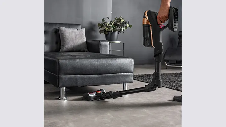 Salon z szarą sofą, rośliną w doniczce i osoba używająca czarnego odkurzacza.
