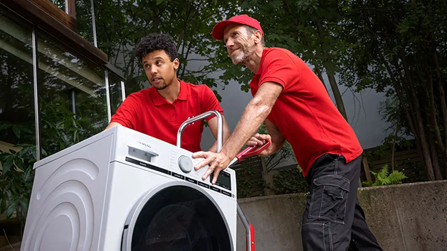 Dwóch mężczyzn w czerwonych koszulkach transportuje białą pralkę na wózku transportowym na zewnątrz budynku.