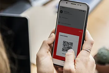 Osoba trzyma biały smartfon z kodem QR na czerwonym tle. W tle widać laptop i biurko.