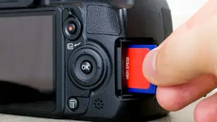 Czarny aparat fotograficzny z ekranem i ręką wkładającą czerwoną kartę pamięci.