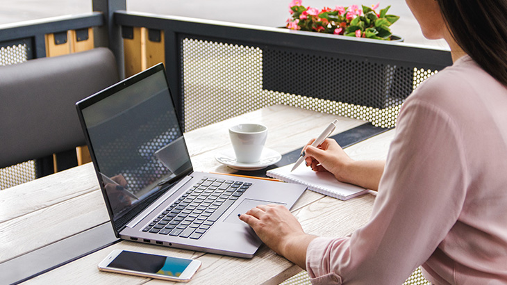 Osoba w różowej koszuli pracuje na laptopie, obok stoi filiżanka i smartfon