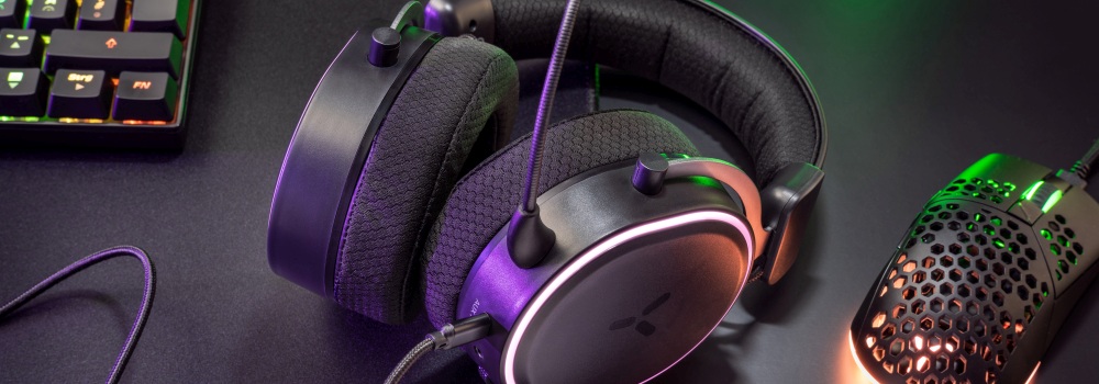 Na ciemnym biurku leżą czarne słuchawki z mikrofonem, klawiatura oraz perforowana mysz z podświetleniem RGB.